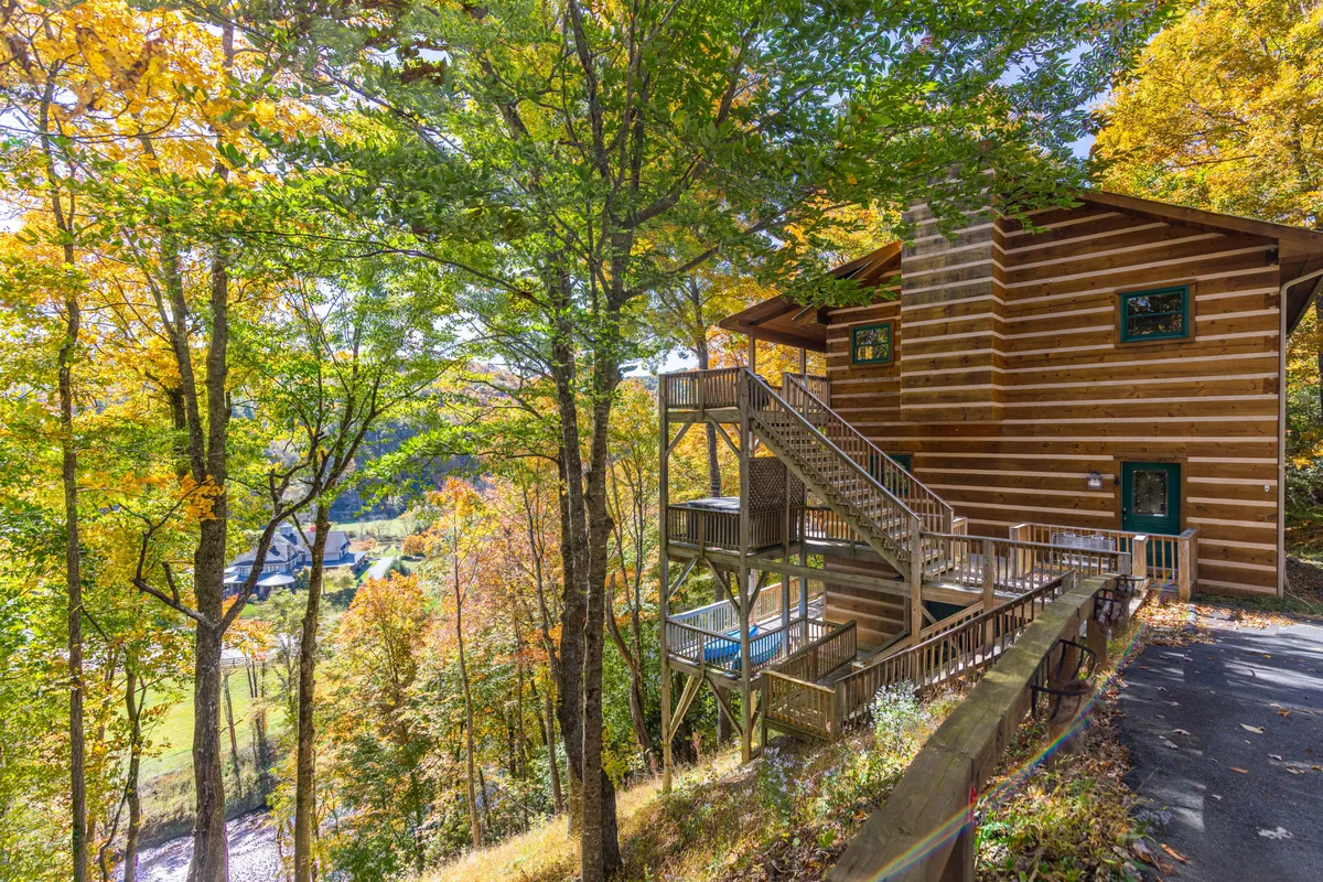 A wide exterior view of the log cabin, highlighting an expansive deck with comfortable seating, surrounded by towering trees and overlooking a breathtaking mountain valley. The image captures serene sunset light, inviting relaxation and connection with the natural setting.
