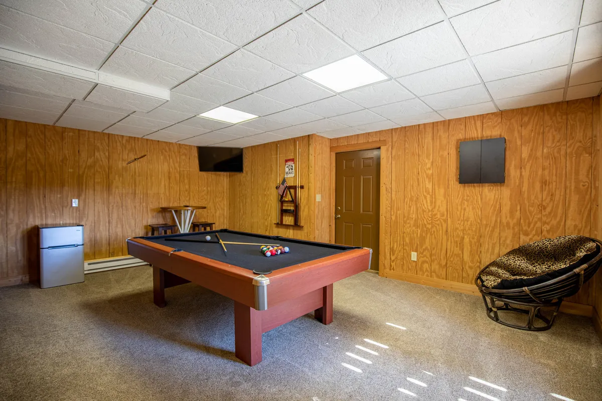 This image showcases a spacious game room featuring a pool table, a reclining chair, a television, a door for deck access, and a mini fridge. The setting is designed for both competitive fun and relaxed socializing, with ample room for guests to gather and enjoy friendly games or movie nights.
