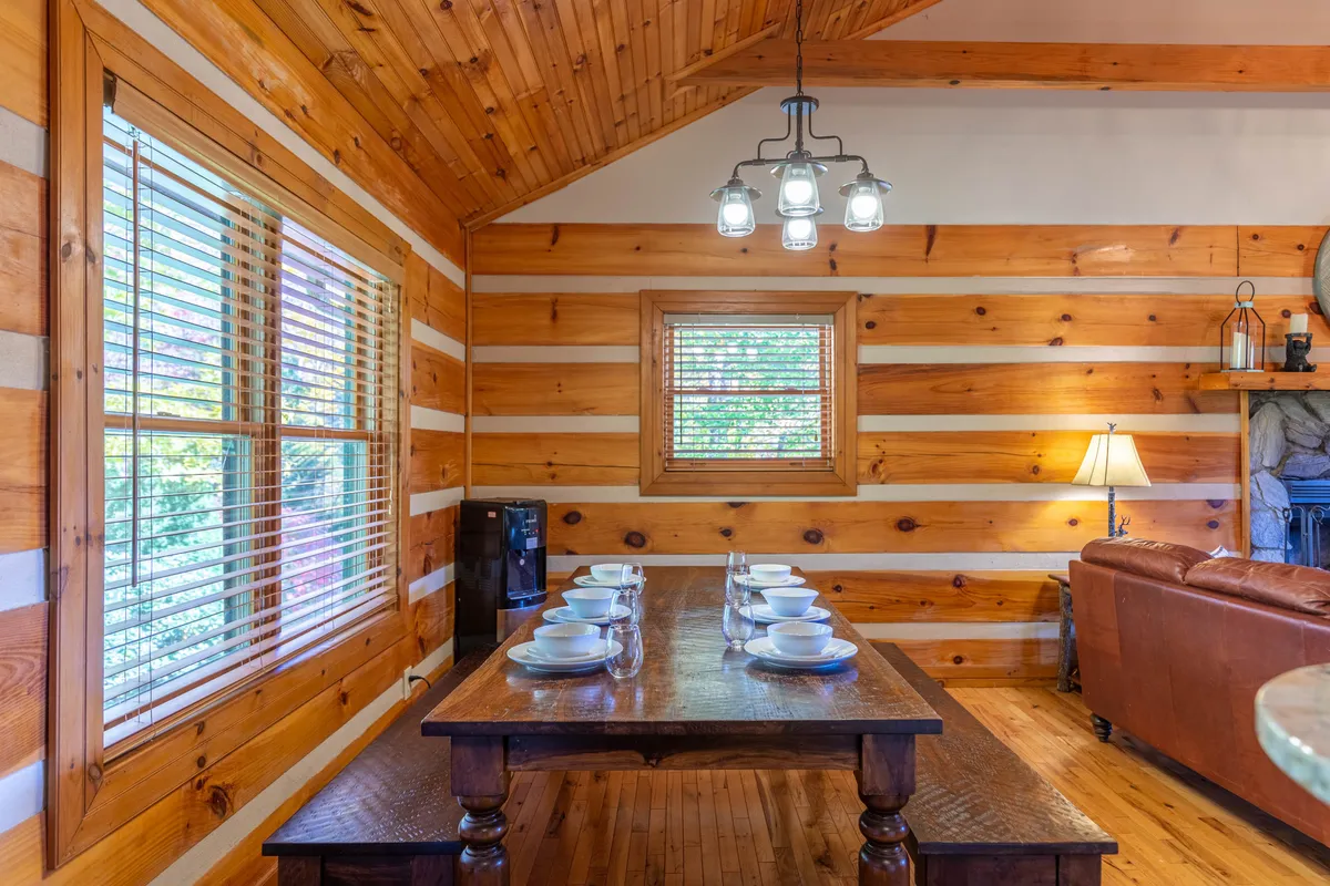 A warm and inviting dining area features a wooden table set near expansive windows with blinds, adjacent to a leather sofa. The space is styled for family gatherings, morning coffee rituals, and game nights, with an overall welcoming and communal vibe.