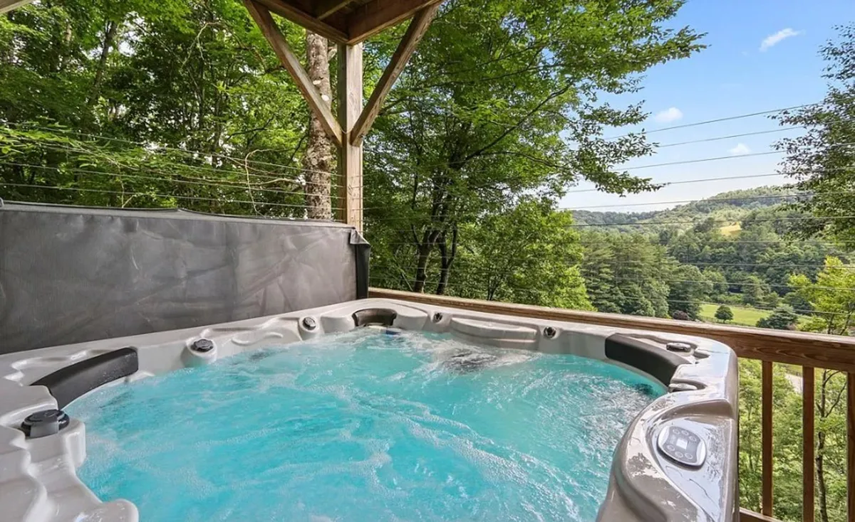 An inviting hot tub set on a private deck, surrounded by natural landscape and open sky. The view extends to rolling hills and forested areas, suggesting a tranquil spot for soaking while immersed in mountain air.