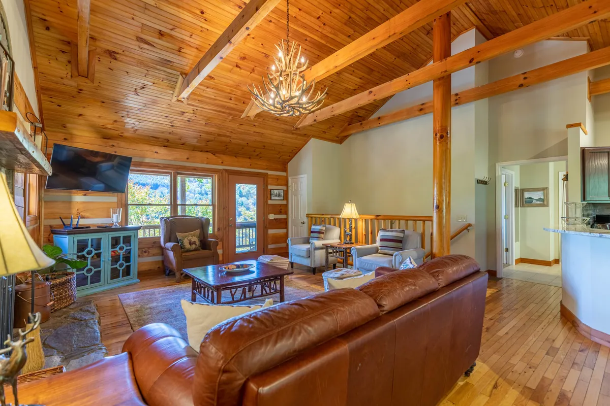 This image presents a cozy, open-concept living area with a large chandelier, exposed wooden beams, a comfortable seating arrangement, a welcoming fireplace, and prominent large windows that frame scenic outdoor views. The space is bathed in natural light and appears perfect for gatherings and relaxation.