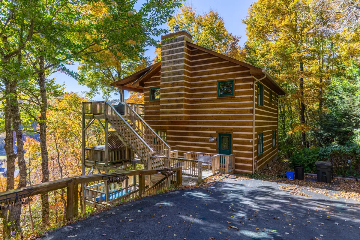 The image showcases a charming log cabin nestled among tall trees, with inviting wooden stairs leading up to a spacious deck. The surrounding greenery creates a peaceful and private setting, perfect for family gatherings, sunrise views, and outdoor barbecues.
