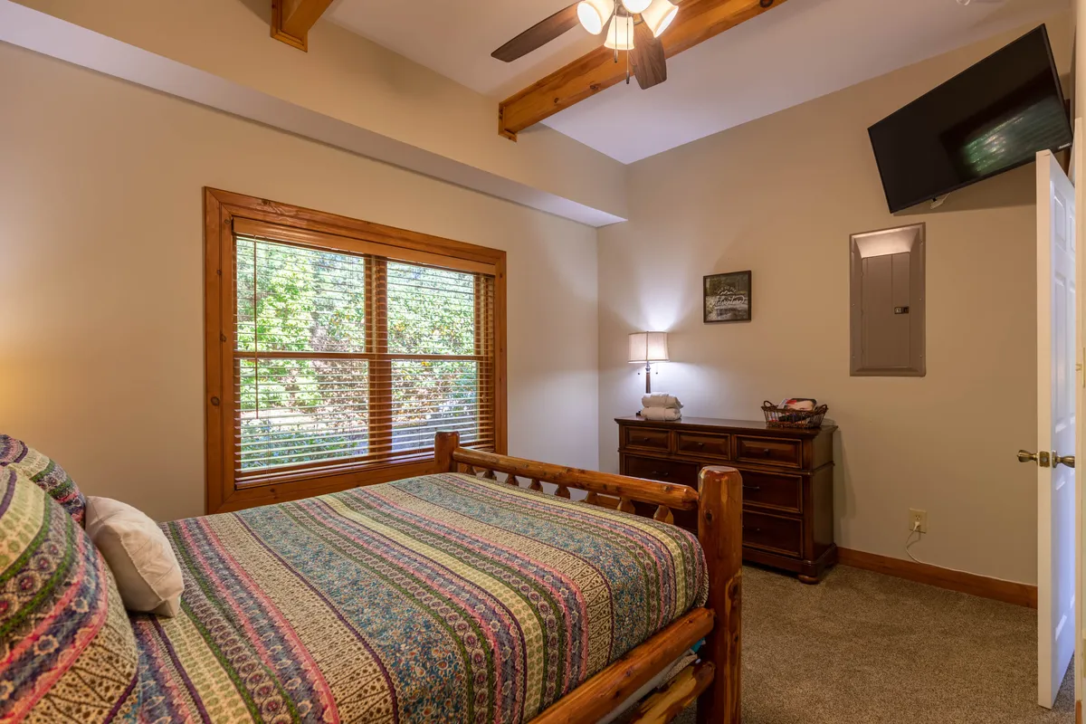 A log bed adorned with patterned bedding is positioned near a window that lets in abundant natural light. A wooden dresser and a table lamp add to the calm, restful mood, suggesting a serene retreat after a day of adventure.