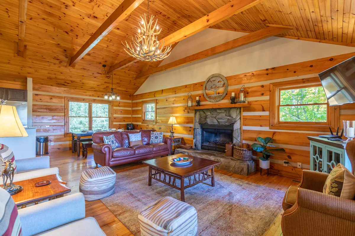 This inviting scene features a crackling wood-burning fireplace, a striking chandelier, plush leather sofas, and a dining area, all under vaulted wood-paneled ceilings. The ambiance is warm and perfect for relaxing, storytelling, or enjoying a cozy evening indoors.