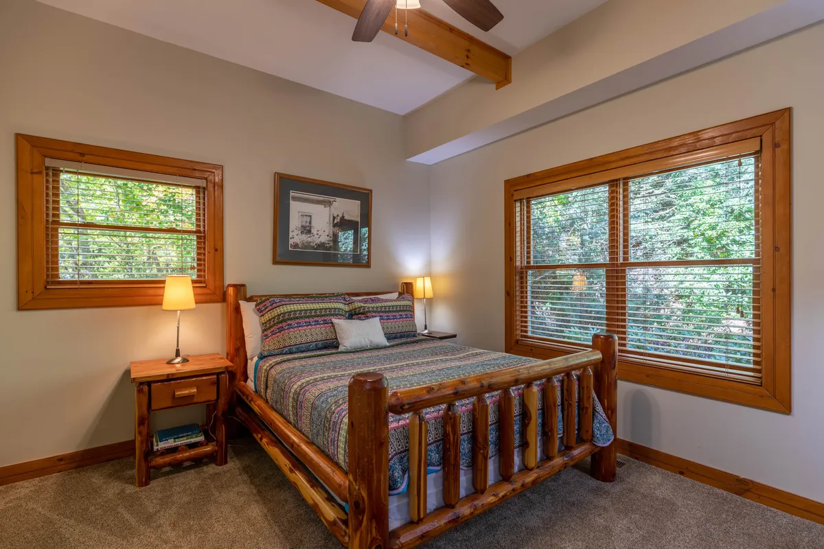 A cozy log bed with a colorful bedspread sits beneath warm lamps, flanked by windows that invite sunlight to filter into the room. The wooden textures and soft lighting create a restful, inviting atmosphere ideal for relaxation and reading.
